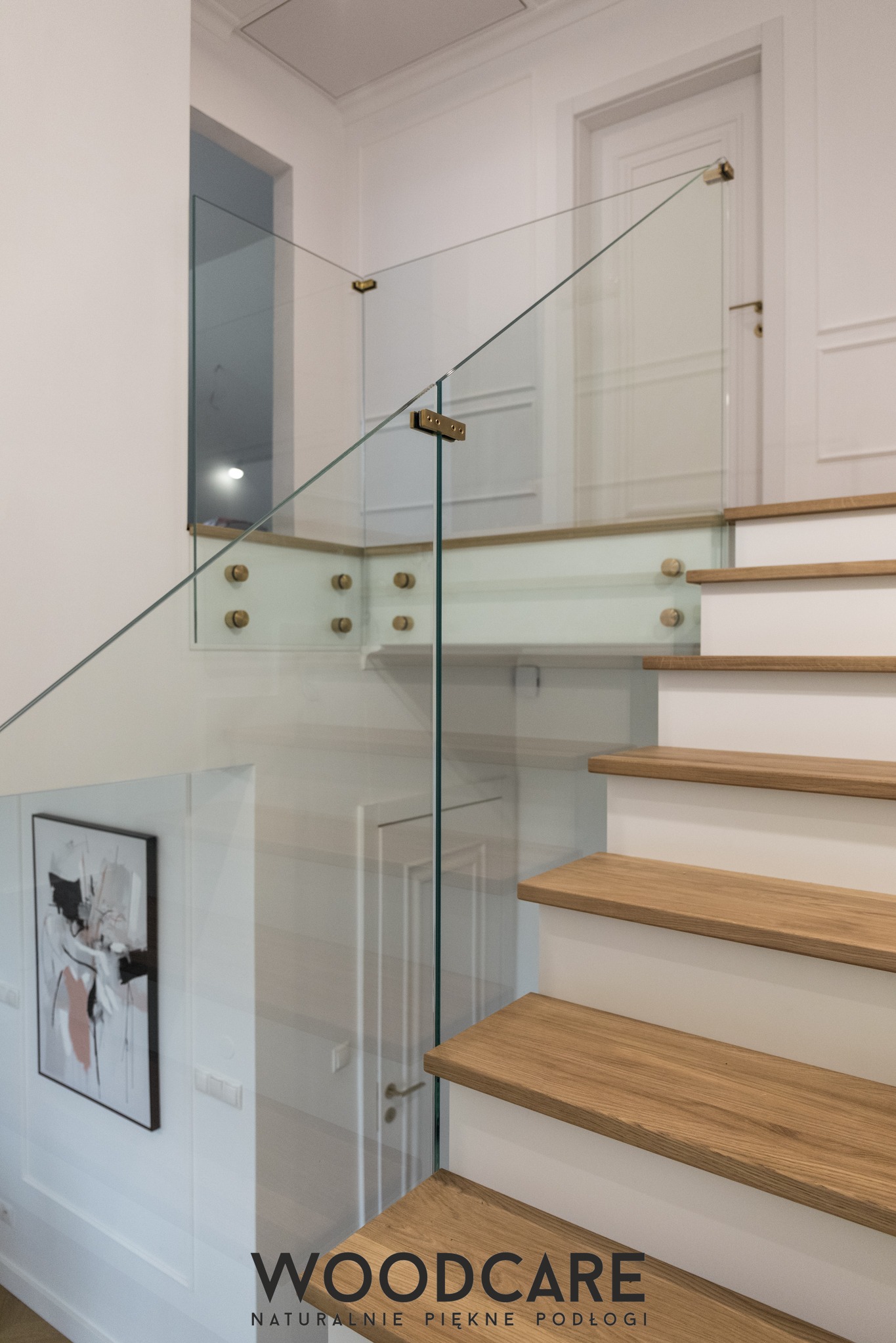 Nowoczesne schody z drewnianymi stopniami i szklaną balustradą, mocowaną na złotych elementach. W tle białe ściany z dekoracyjnymi panelami i obraz.