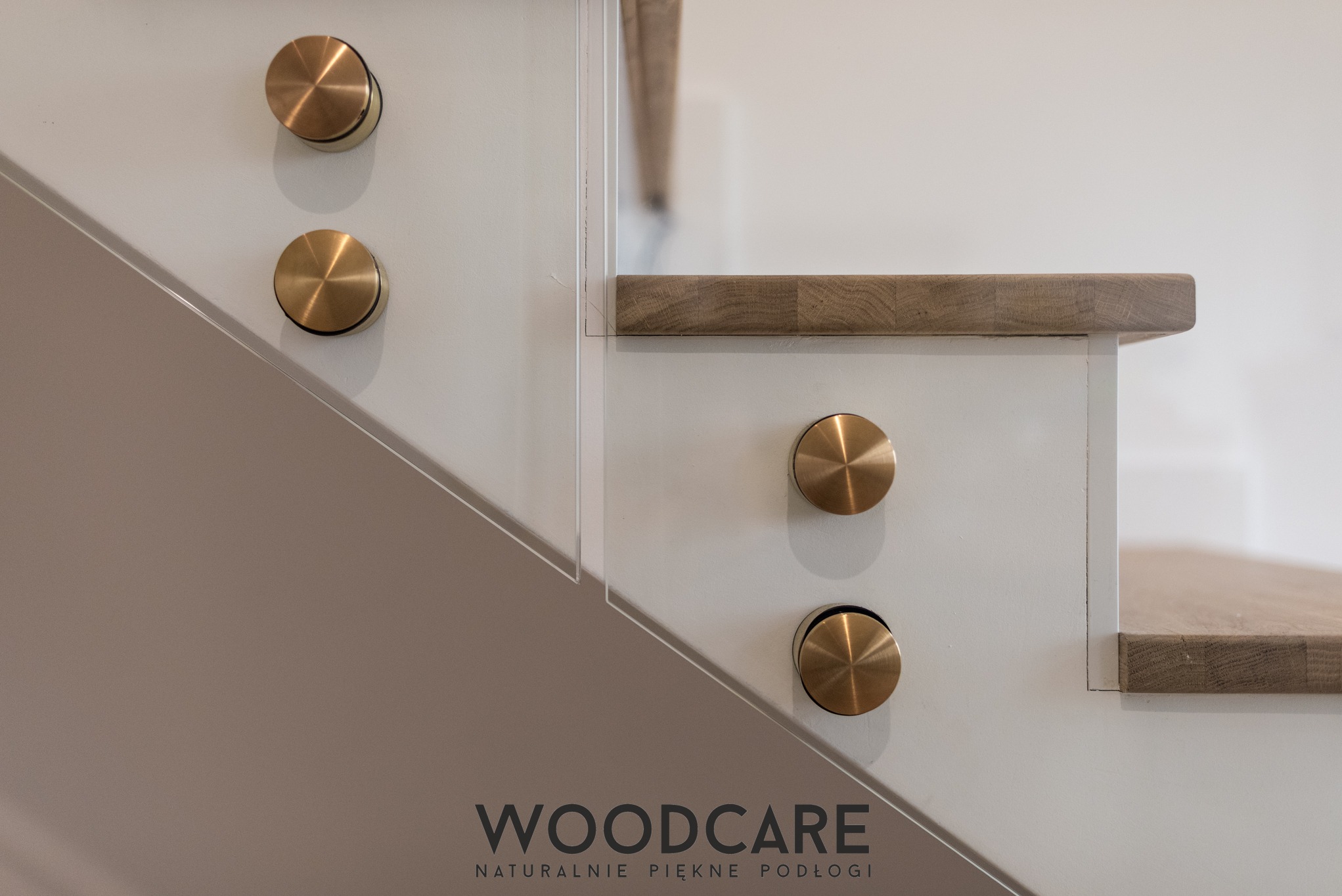 Detal drewnianych schodów z balustradą ze szkła, mocowaną złotymi uchwytami. Widoczny fragment stopnia i boku schodów. Minimalistyczny design.