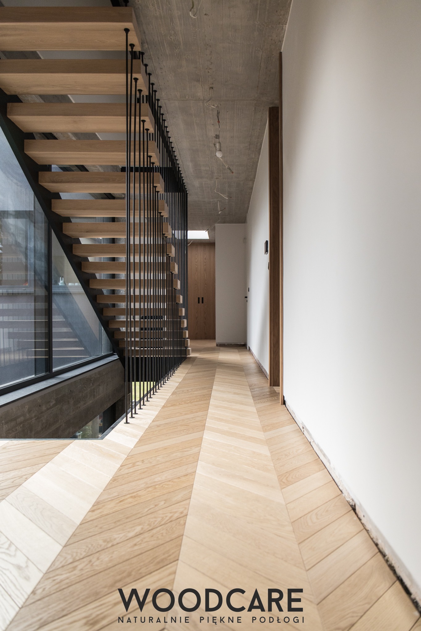 Nowoczesne schody drewniane z czarną balustradą i parkietem ułożonym we wzór jodełki w jasnym, minimalistycznym wnętrzu z surowym betonowym sufitem.
