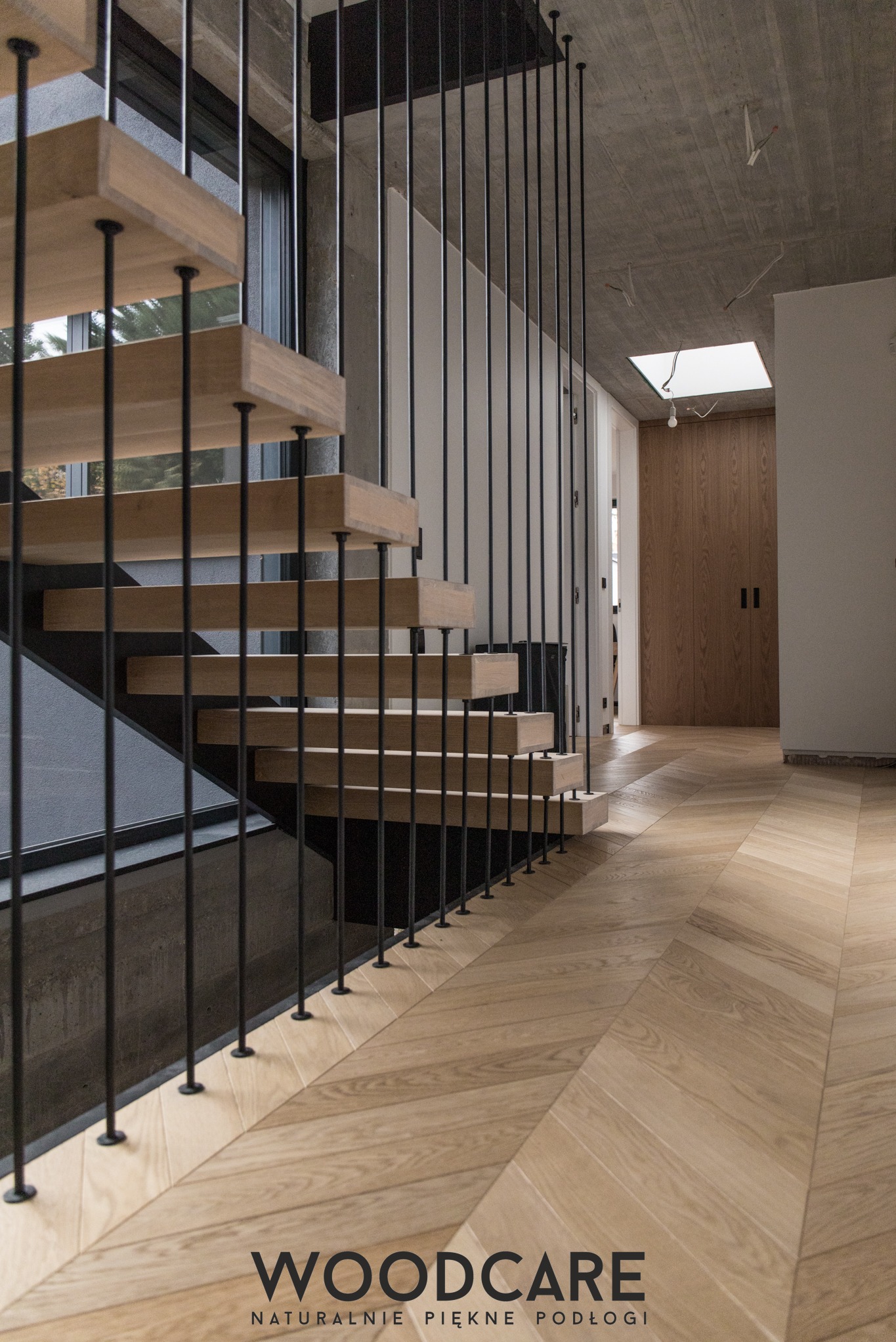 Nowoczesne schody z drewnianymi stopniami i czarną, metalową balustradą. Parkiet ułożony w jodełkę. Widoczne surowe, betonowe elementy i okno. Minimalistyczny design.