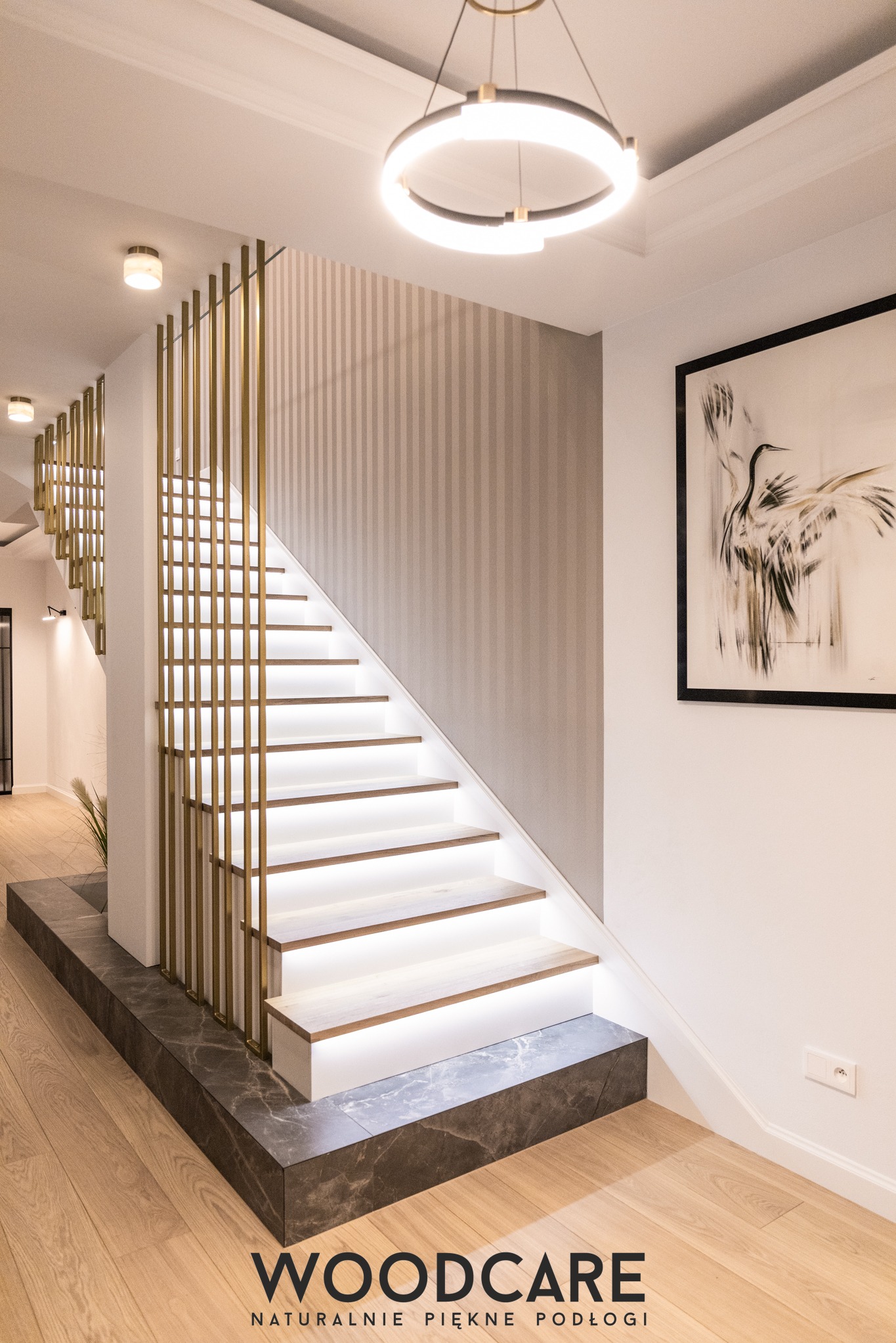 Nowoczesne schody z podświetleniem LED i złotą balustradą w eleganckim wnętrzu. Widoczny fragment parkietu i obraz na ścianie. Minimalistyczny design.