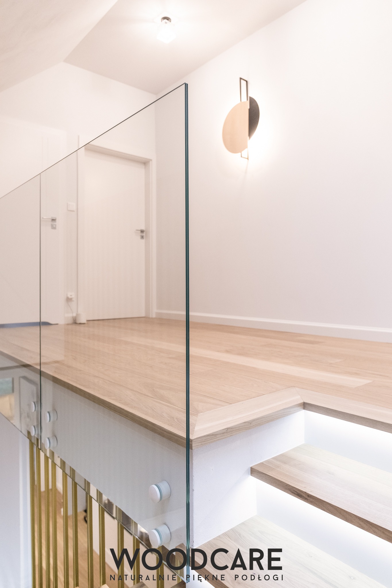 Jasne drewniane schody z podświetleniem LED i szklaną balustradą. Widoczne złote elementy balustrady i fragment parkietu. Minimalistyczny design, nowoczesne oświetlenie.