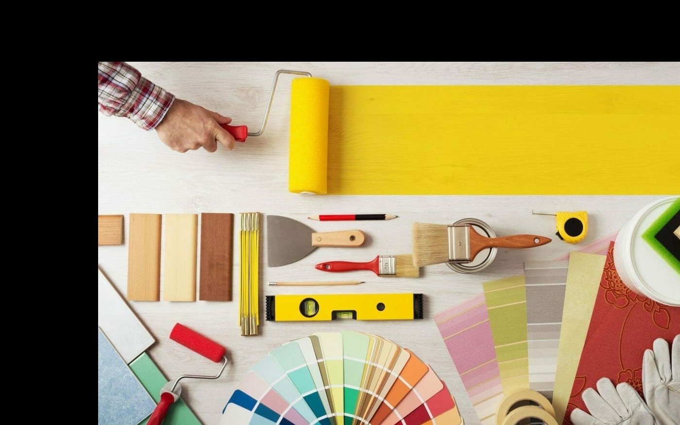 Widok z góry na narzędzia malarskie i próbki kolorów na białym drewnianym tle, w tym wałek malarski w akcji, pędzle, szpachelka, poziomica, miarka, paleta kolorów i próbki tapet.
