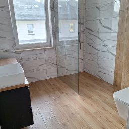 INFRARED HOME SP. z o.o. - Wykończona łazienka z kabiną prysznicową typu walk-in, umywalką nablatową i toaletą. Płytki imitujące drewno na podłodze i marmur na ścianach. Widok na okno.
