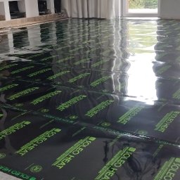 INFRARED HOME SP. z o.o. - Rozłożone czarne maty grzewcze SOLEO HEAT na podłodze w jasnym pomieszczeniu z oknami i kominkiem. Widoczne refleksy światła na folii ochronnej.