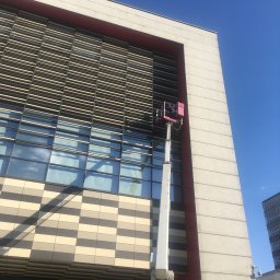 GODT VALG Jarosław Kreciński - Mycie okien na wysokościowcu przy użyciu podnośnika koszowego z czerwoną platformą, widoczny fragment białej furgonetki w dolnej części kadru.