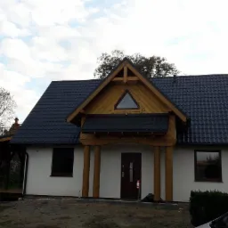 Dom jednorodzinny z nowym, grafitowym dachem z blachodachówki, drewnianą konstrukcją nad wejściem i trójkątnym oknem na szczycie, widok z poziomu gruntu, częściowo zasłonięty przez samochód...