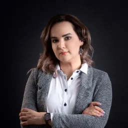 Portret kobiety w szarej marynarce i białej koszuli, z założonymi rękami, na ciemnym tle. Widoczne kolczyki i zegarek.