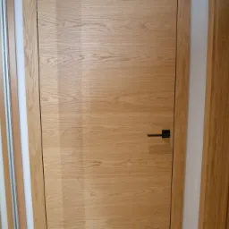 Zamknięte, drewniane drzwi wewnętrzne z czarną klamką, widoczne w białej ościeżnicy.