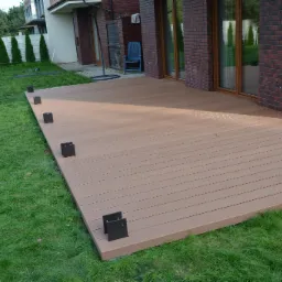 Nowo zbudowany taras z desek kompozytowych w kolorze jasnego brązu, z widocznymi czarnymi wspornikami, otoczony zielonym trawnikiem i budynkiem z ceglanymi elementami.