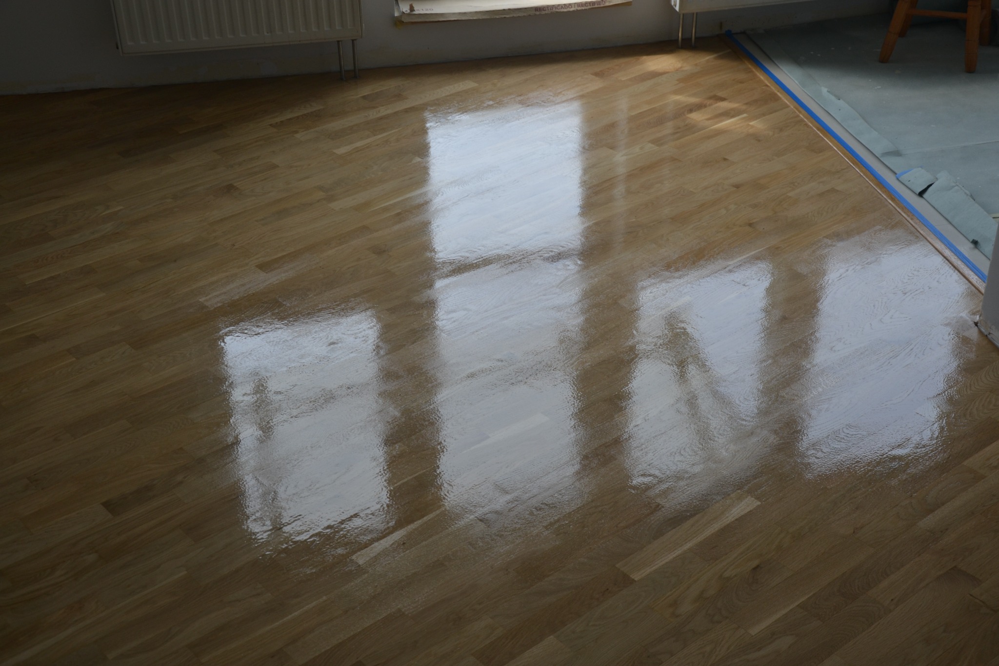 Błyszcząca, świeżo polakierowana podłoga parkietowa z widocznymi odbiciami okien, zabezpieczona niebieską taśmą malarską przy krawędzi ściany.