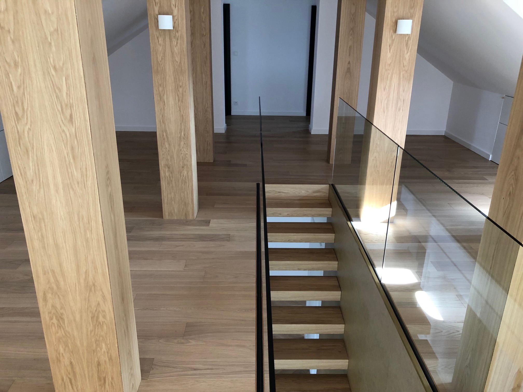 Jasne drewniane schody z czarnymi poręczami i szklaną balustradą, widok z góry na poddasze z drewnianą podłogą i kolumnami.