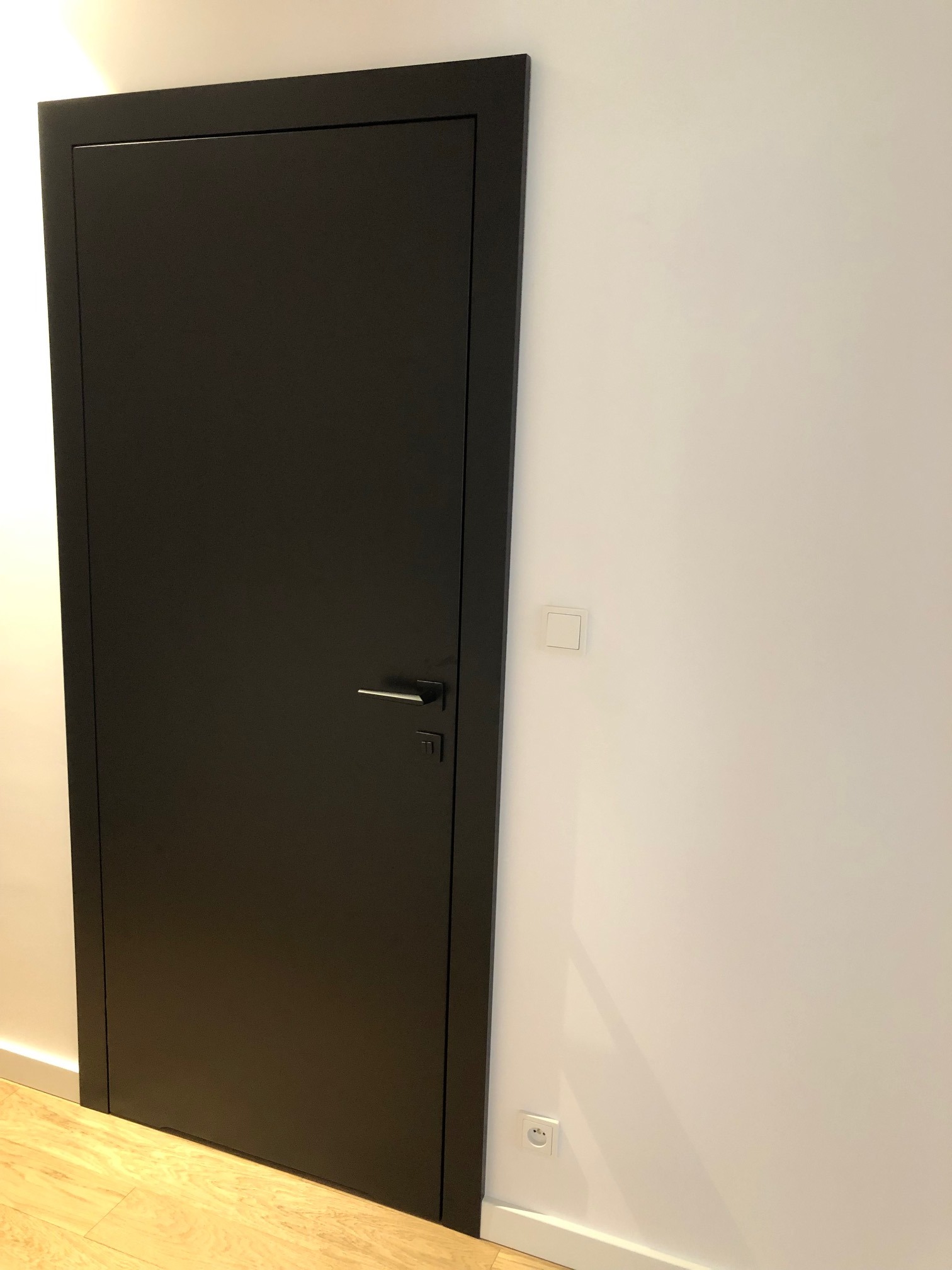 Zamknięte, czarne drzwi wewnętrzne z metalową klamką na tle jasnej ściany z gniazdkiem i włącznikiem światła, widoczna podłoga z paneli.