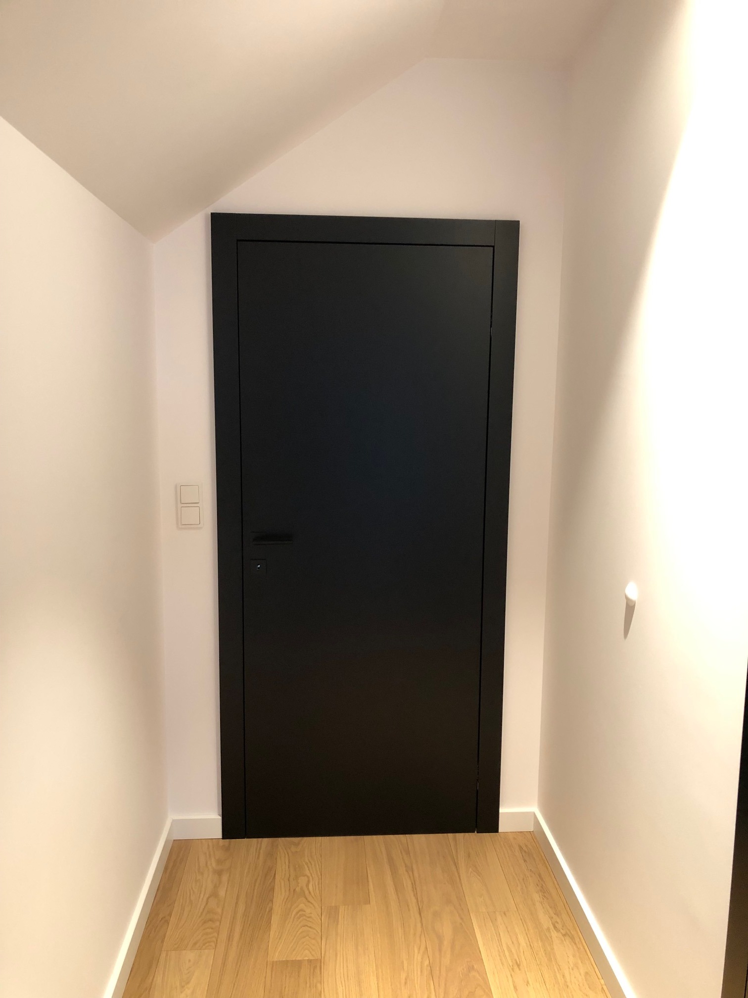 Czarna, minimalistyczna wewnętrzna drzwi w białej wnęce, z jasną drewnianą podłogą, widoczne włączniki światła.