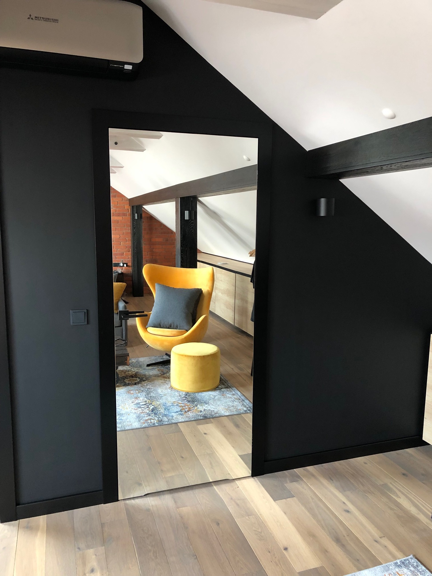 Widok na odnowione poddasze z żółtym fotelem i pufą, przez lustrzane drzwi w czarnej ramie, parkiet w jasnym drewnie, czarne ściany, ceglana ściana w tle.