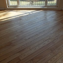 Król Parkietu - Świeżo wycyklinowana podłoga z jasnego drewna, widoczne słońce padające przez okno z metalową kratą, ukazujące unikalny wzór słojów drewna, wykończona listwa przypodłogowa.