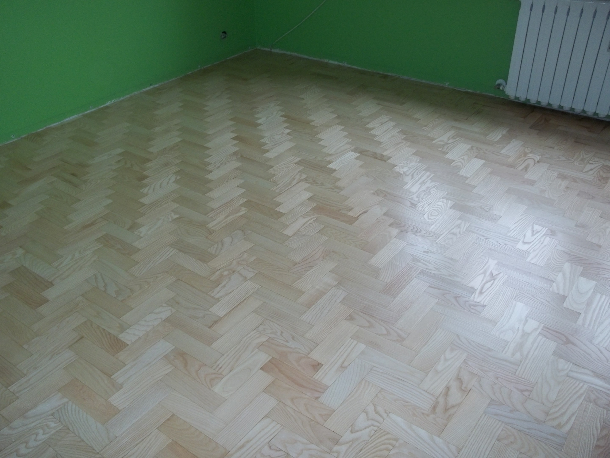 Jasny parkiet ułożony we wzór jodełki francuskiej w pokoju z zielonymi ścianami i białym grzejnikiem.
