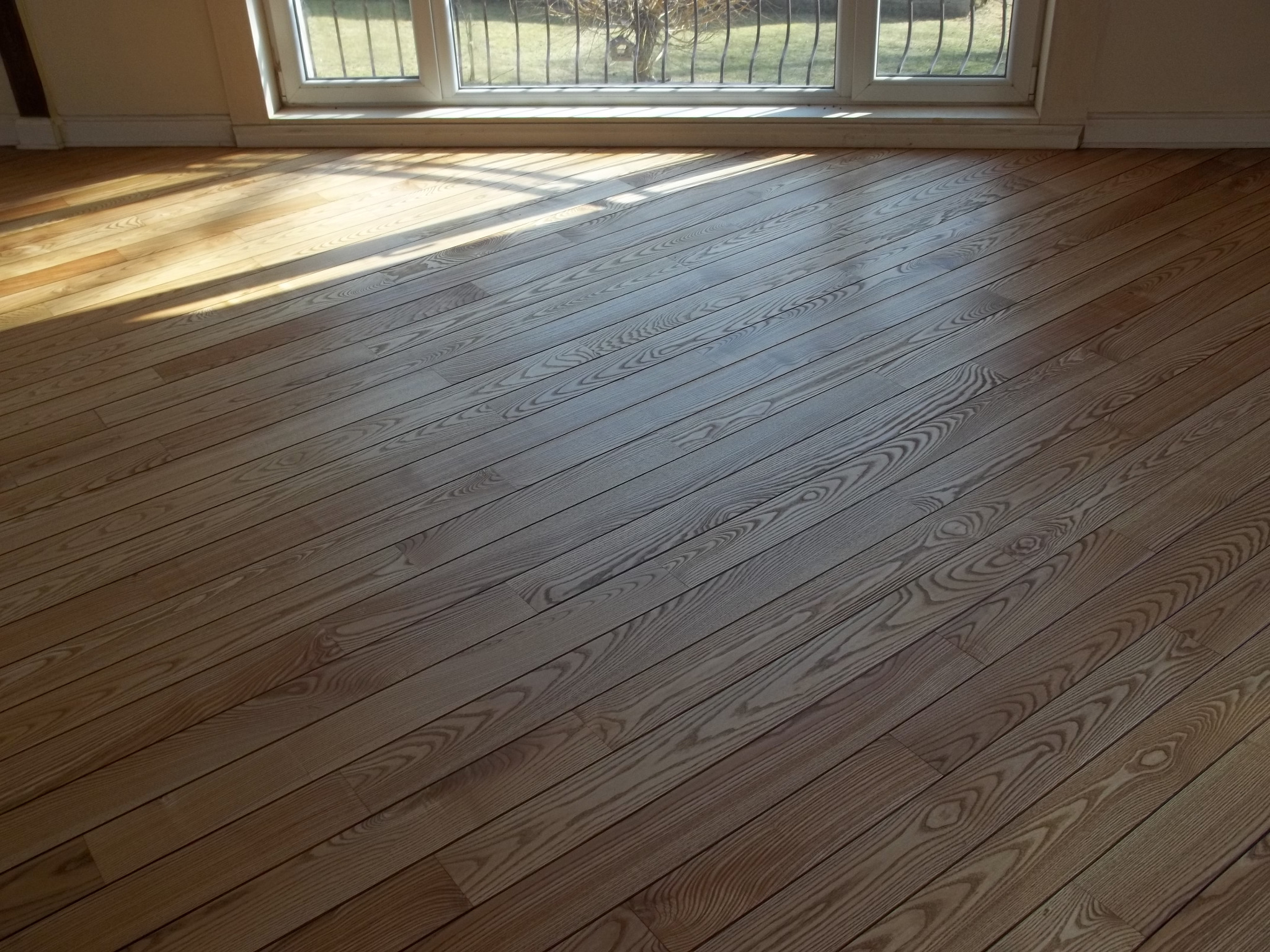 Świeżo wycyklinowana podłoga z jasnego drewna, widoczne słońce padające przez okno z metalową kratą, ukazujące unikalny wzór słojów drewna, wykończona listwa przypodłogowa.