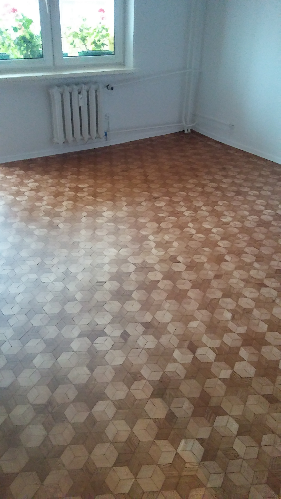 Ułożony parkiet w geometryczny wzór kostki 3D w pustym pokoju z białymi ścianami, grzejnikiem i oknem z zielenią na parapecie.