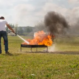 Osoba w białej koszulce gasi płonący ogień w metalowej konstrukcji na trawiastym terenie, używając węża gaśniczego, z widocznym czarnym dymem unoszącym się w g&oacute;rę.