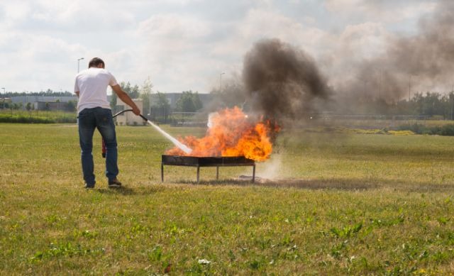 Osoba w białej koszulce gasi płonący ogień w metalowej konstrukcji na trawiastym terenie, używając węża gaśniczego, z widocznym czarnym dymem unoszącym się w górę.