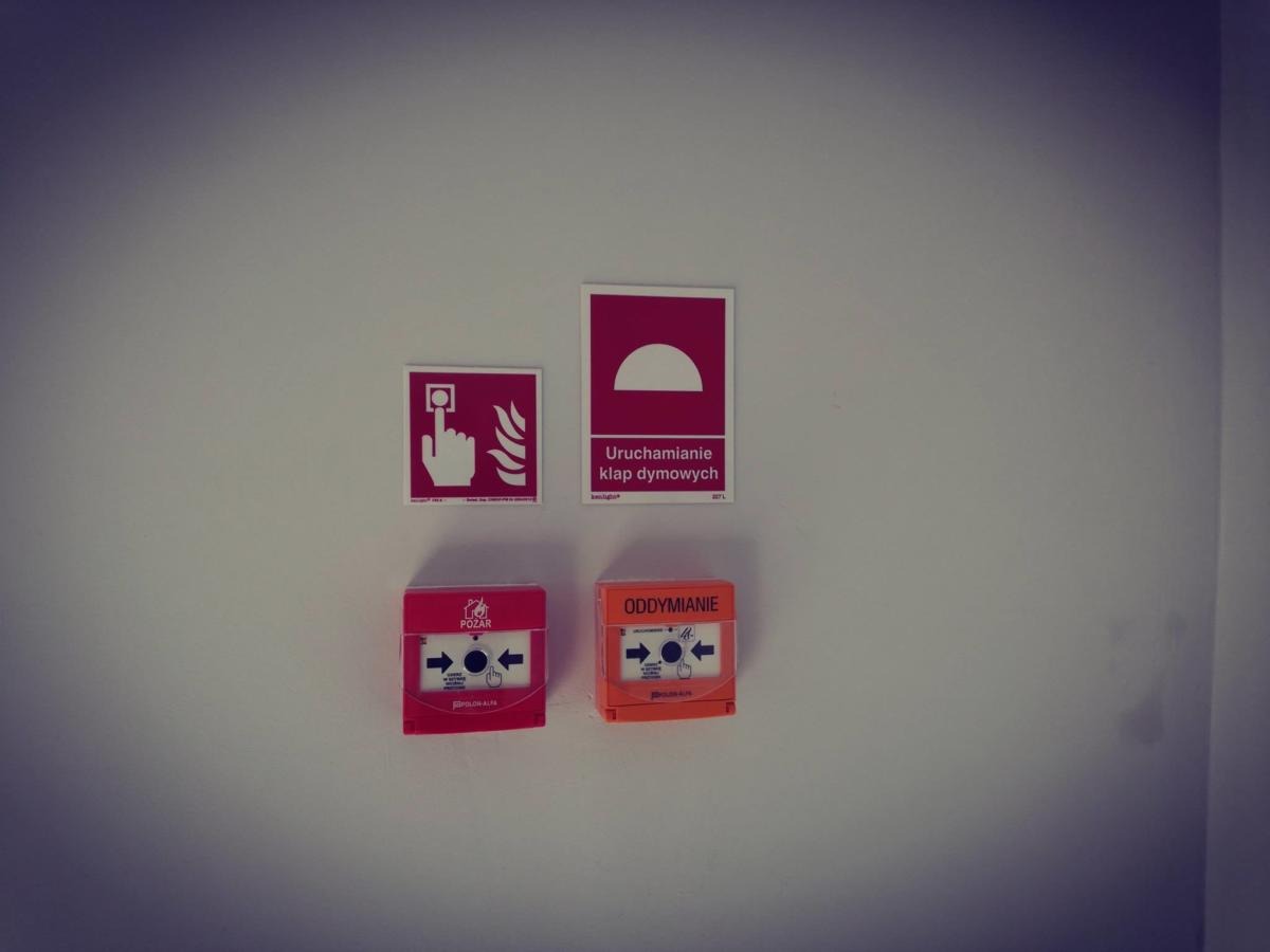 Zestaw znaków i przycisków alarmowych (pożar i oddymianie) zamontowanych na jasnej ścianie wewnątrz budynku.