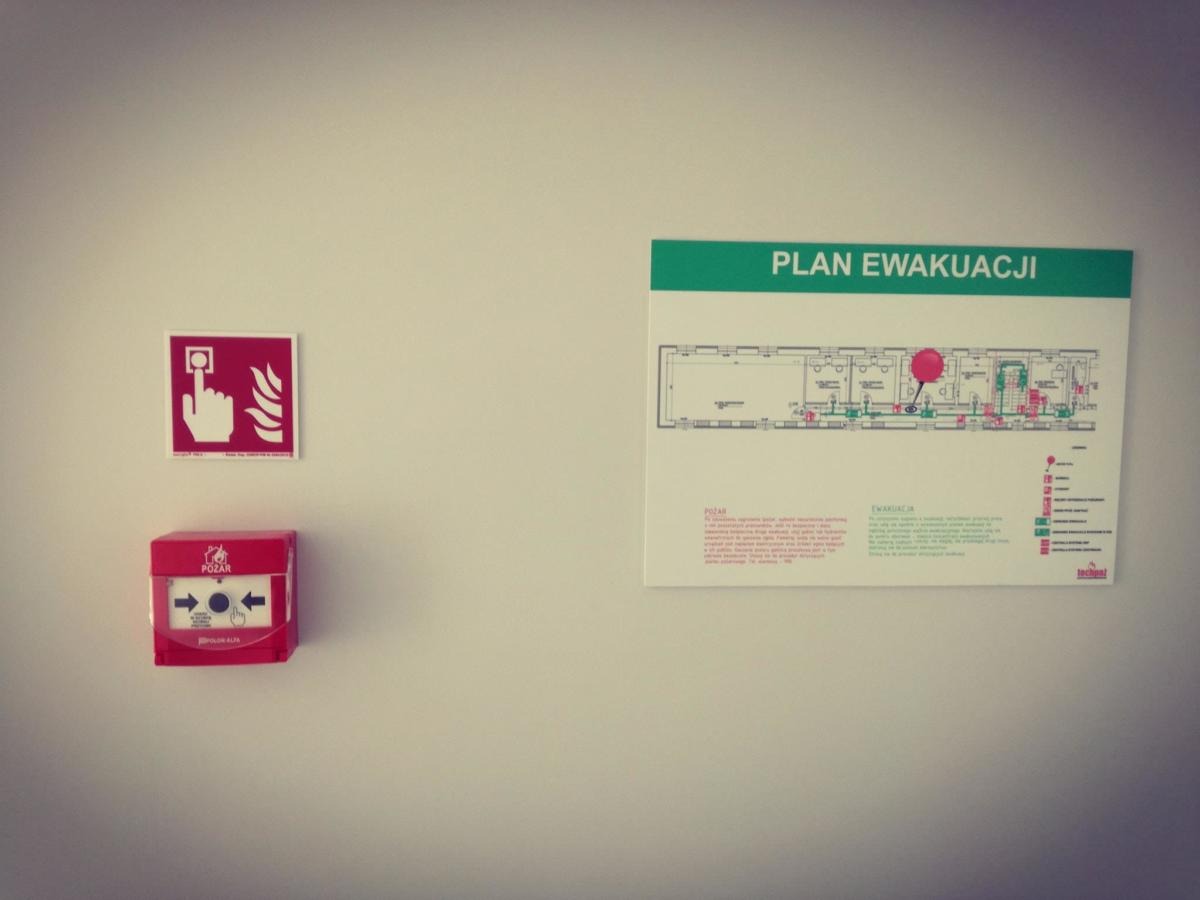 Na jasnej ścianie wisi czerwony przycisk alarmowy POŻAR, znak z dłonią naciskającą przycisk oraz biała tablica z planem ewakuacji z zaznaczoną drogą ucieczki i legendą.
