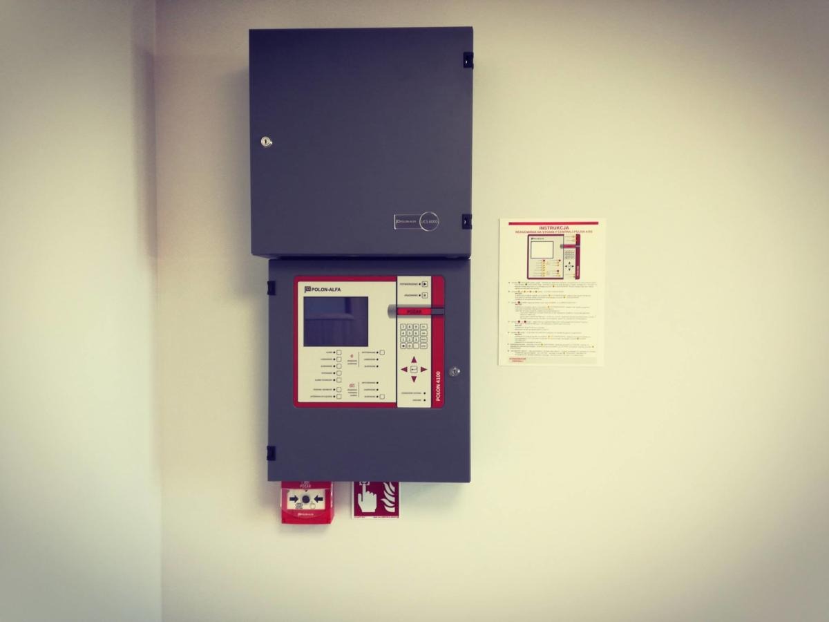 Szara szafka sterownicza systemu alarmowego Polon-Alfa 4100 z czerwonym przyciskiem alarmowym i instrukcją obsługi na białej ścianie.