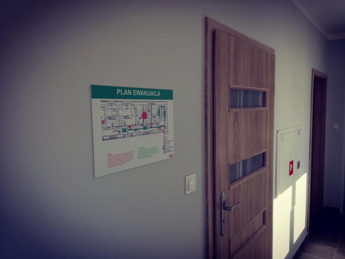 Plan ewakuacji umieszczony na szarej ścianie obok drewnianych drzwi z przeszkleniami i szafki hydrantowej.