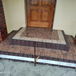 Ułożone schody wejściowe z kostki brukowej w odcieniach brązu i beżu, z widocznymi stalowymi szpilkami stabilizującymi obrzeże, prowadzące do drewnianych drzwi.