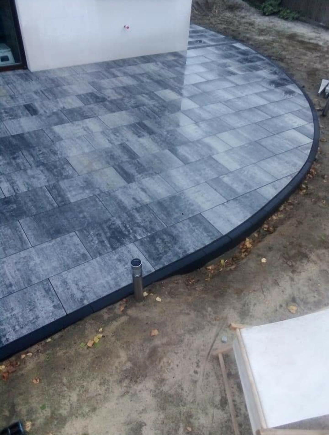 Półokrągły taras wyłożony szarą kostką brukową z ciemną obwódką, widoczny słupek oświetleniowy i fragment leżaka.