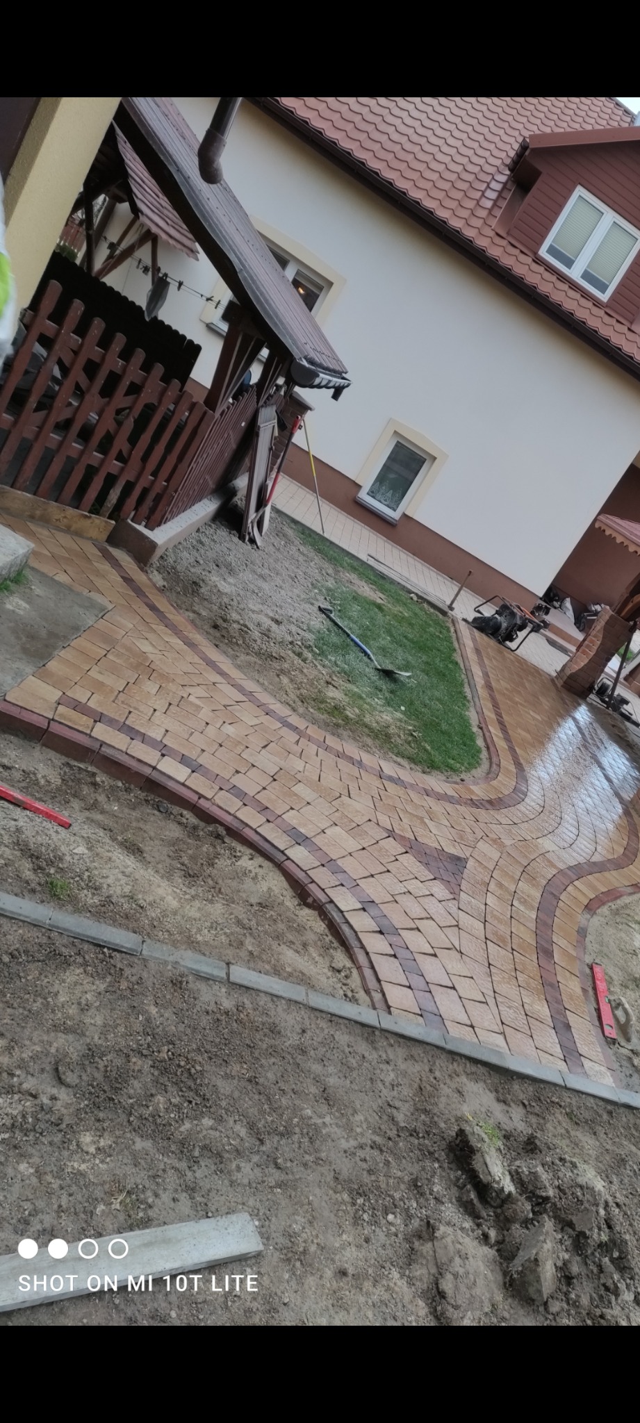 Kaskadowa ścieżka z kostki brukowej w odcieniach beżu i brązu, otoczona ziemią i fragmentami trawnika przed domem z drewnianą altaną.