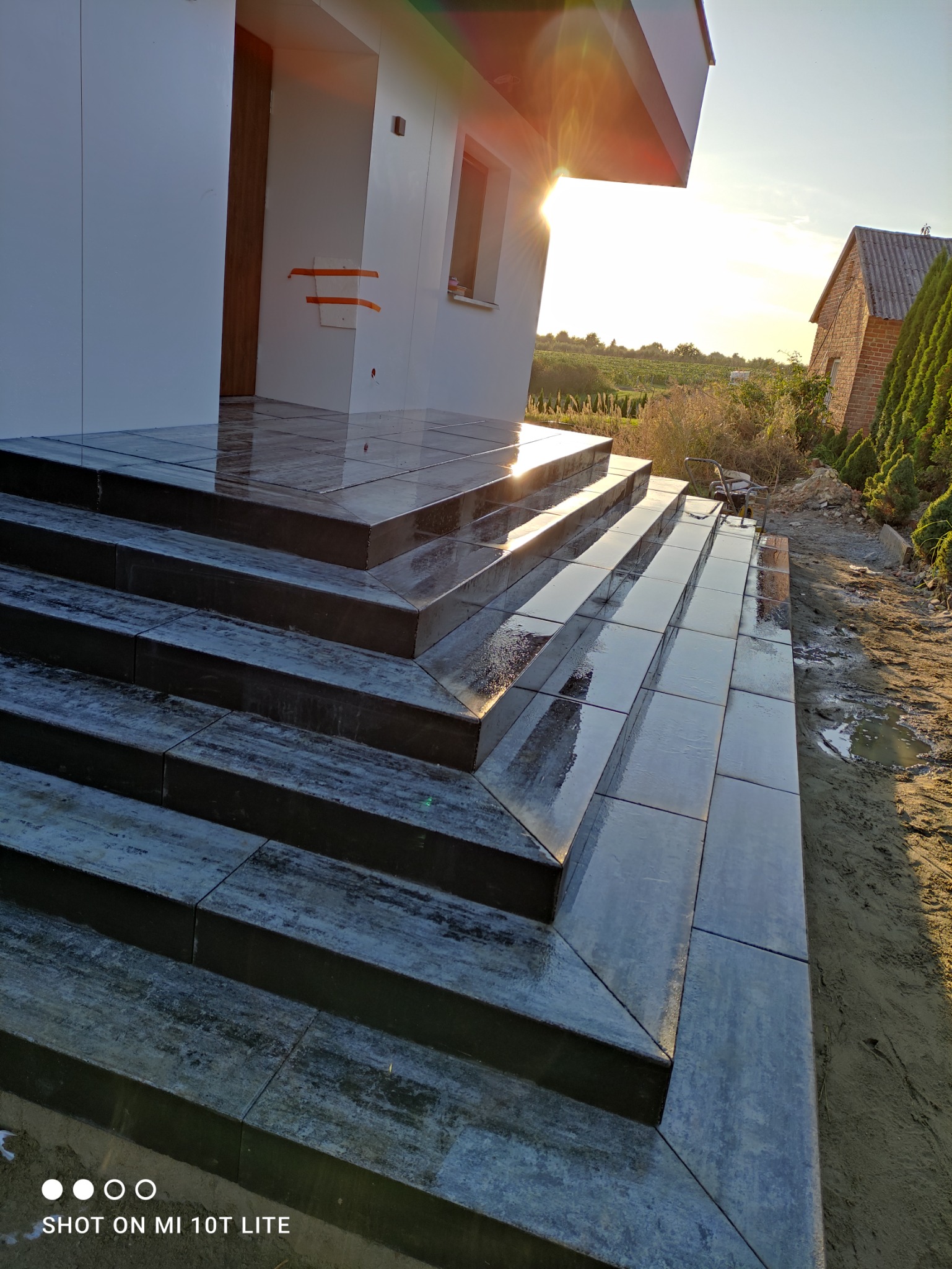 Ułożone, ciemnoszare płytki na schodach wejściowych do nowoczesnego budynku, odbijające promienie zachodzącego słońca. W tle widoczne zabudowania i zieleń.