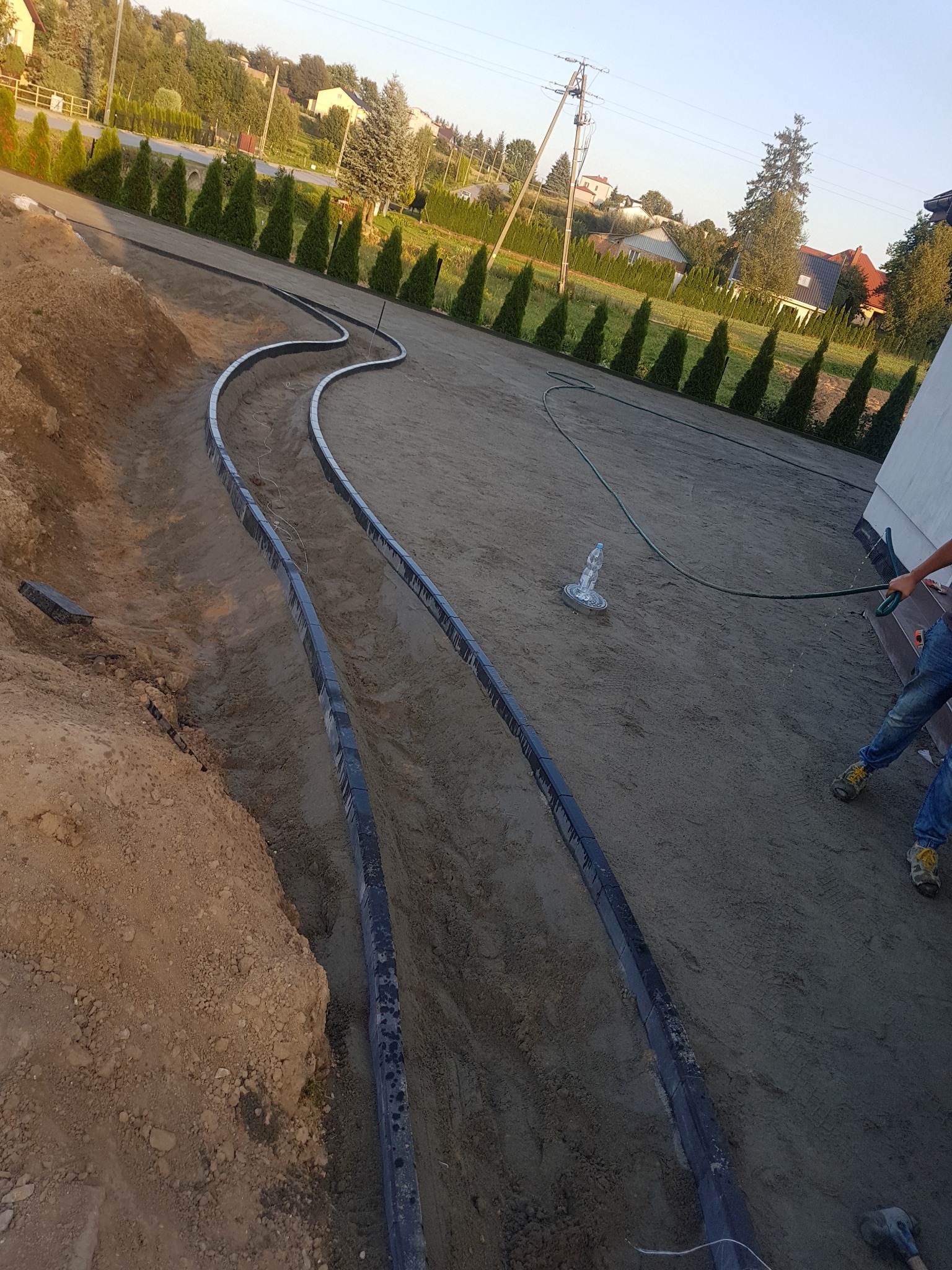 Przygotowanie terenu pod ścieżkę z kostki brukowej: wykopane koryto z ułożonymi obrzeżami, teren wyrównywany wodą z węża, w tle rząd tuj i domy.