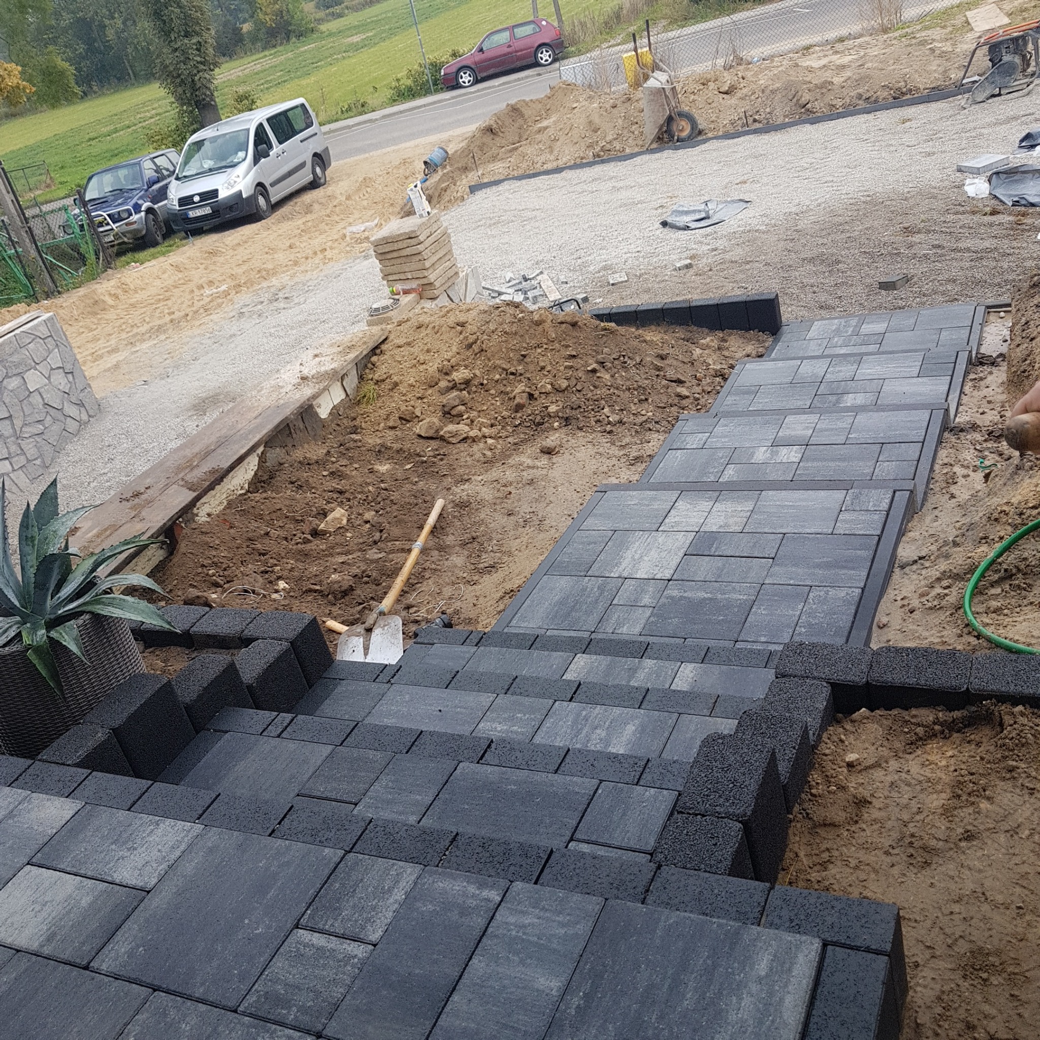 Budowa schodów zewnętrznych z szarych kostek brukowych na nierównym terenie, widoczna łopata wbita w ziemię i donica z rośliną.