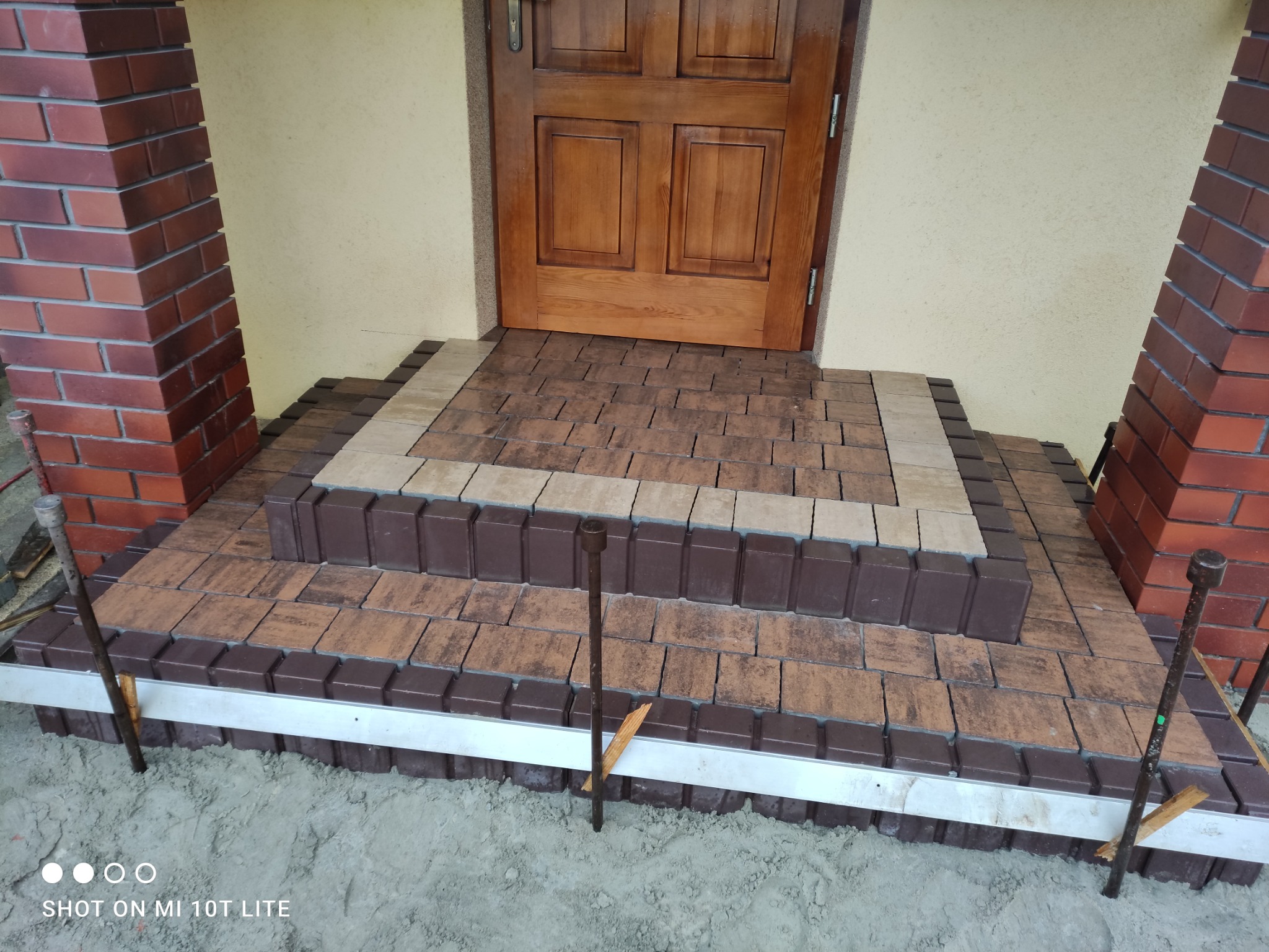 Ułożone schody wejściowe z kostki brukowej w odcieniach brązu i beżu, z widocznymi stalowymi szpilkami stabilizującymi obrzeże, prowadzące do drewnianych drzwi.