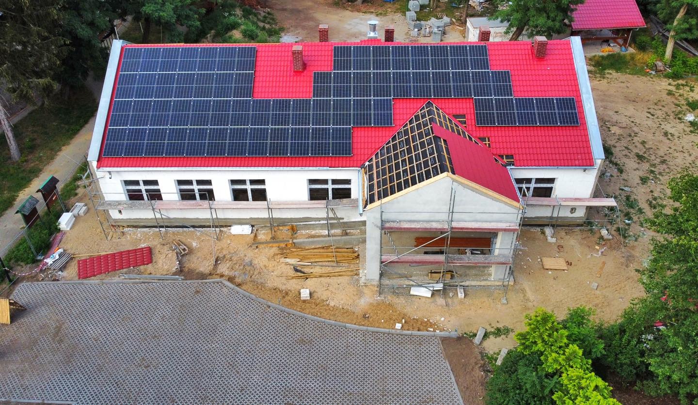 Widok z góry na budynek z czerwonym dachem, na którym zamontowane są panele słoneczne. Do budynku dobudowywana jest nowa część z widoczną konstrukcją dachu i rusztowaniami.