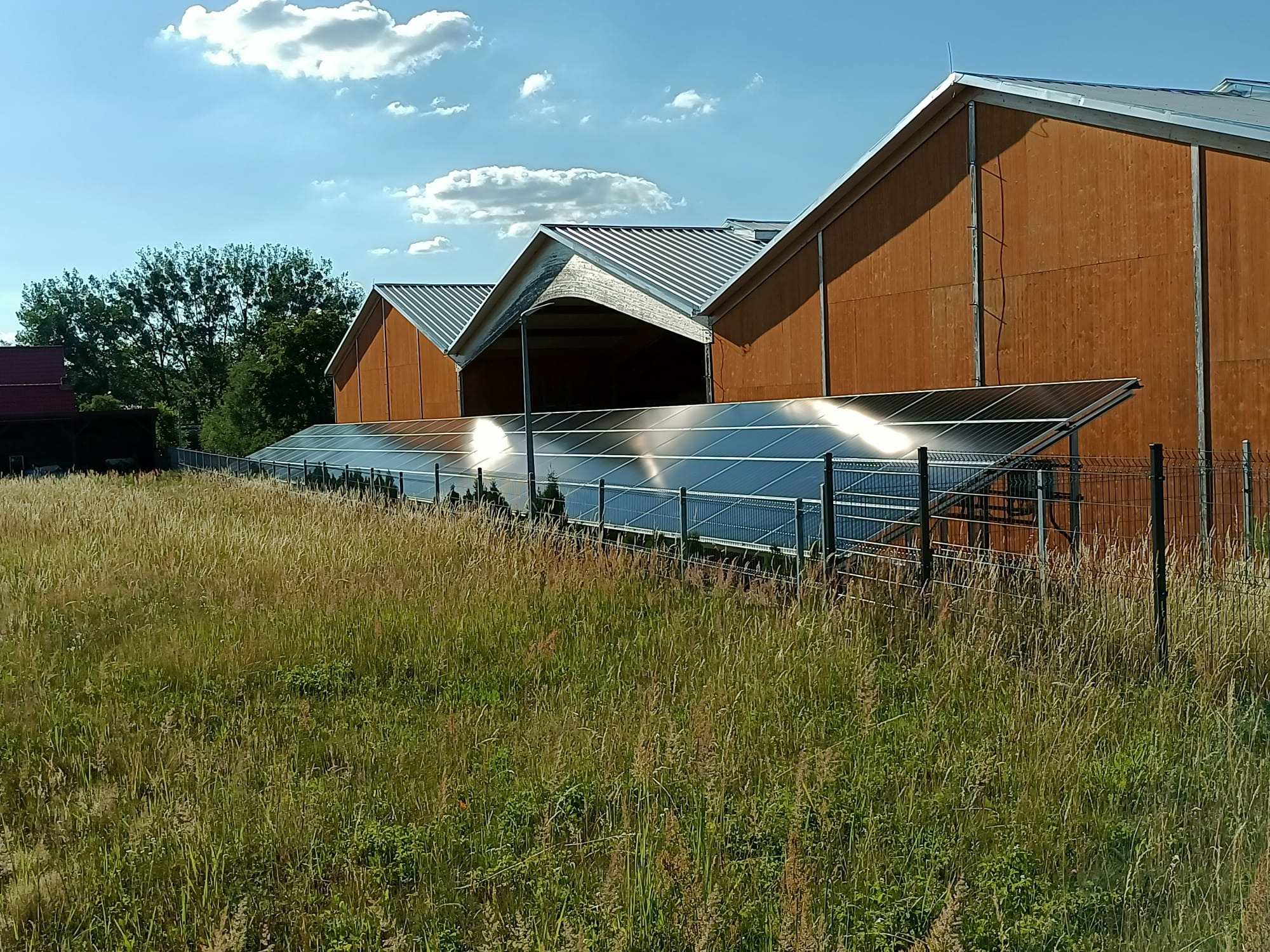 Instalacja paneli słonecznych zamontowana na gruncie, przed drewnianym budynkiem gospodarczym, otoczona wysoką trawą i ogrodzeniem z siatki.