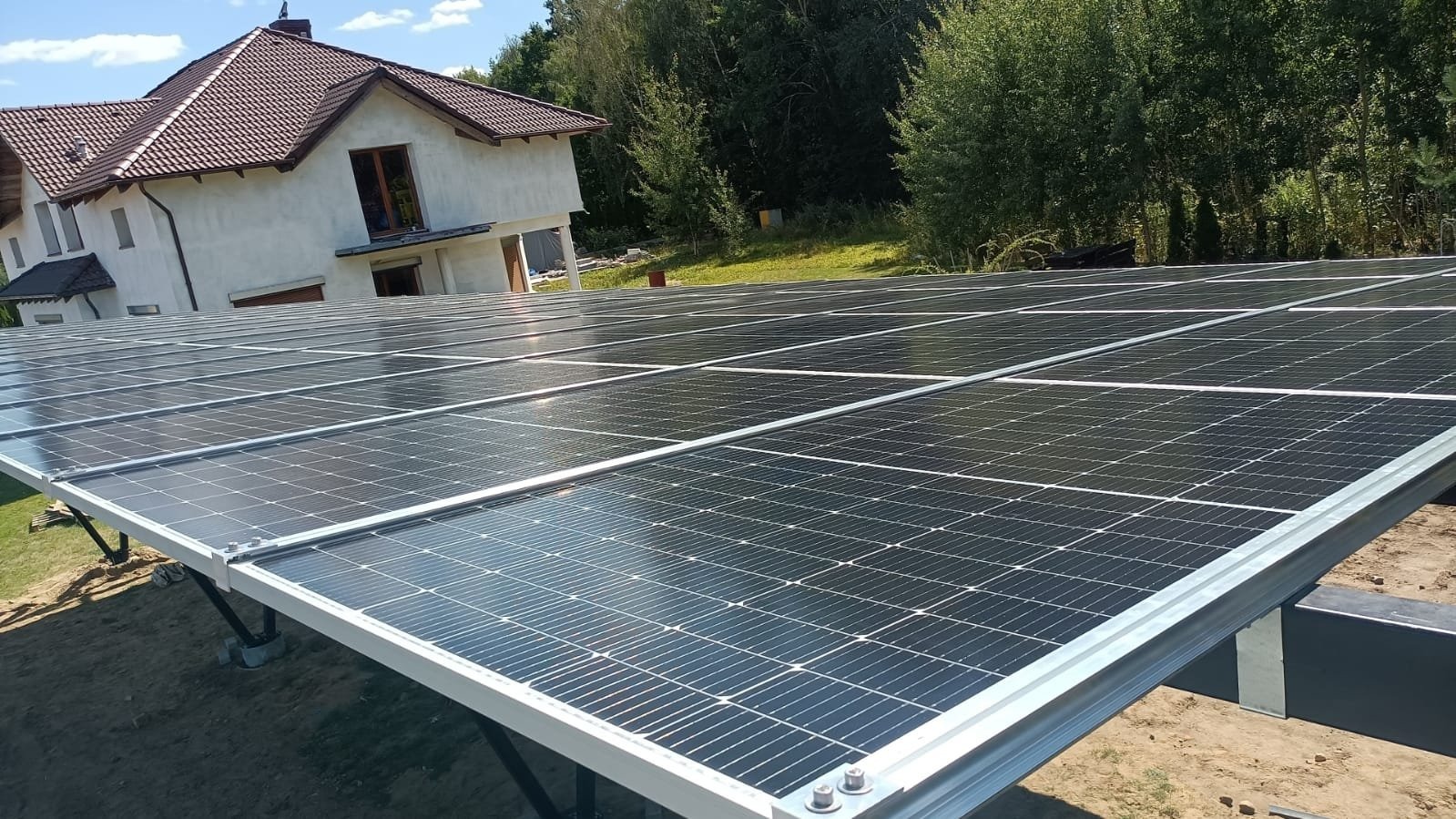 Instalacja paneli słonecznych na konstrukcji naziemnej przed domem jednorodzinnym, widok na panele z perspektywy niskiego kąta.