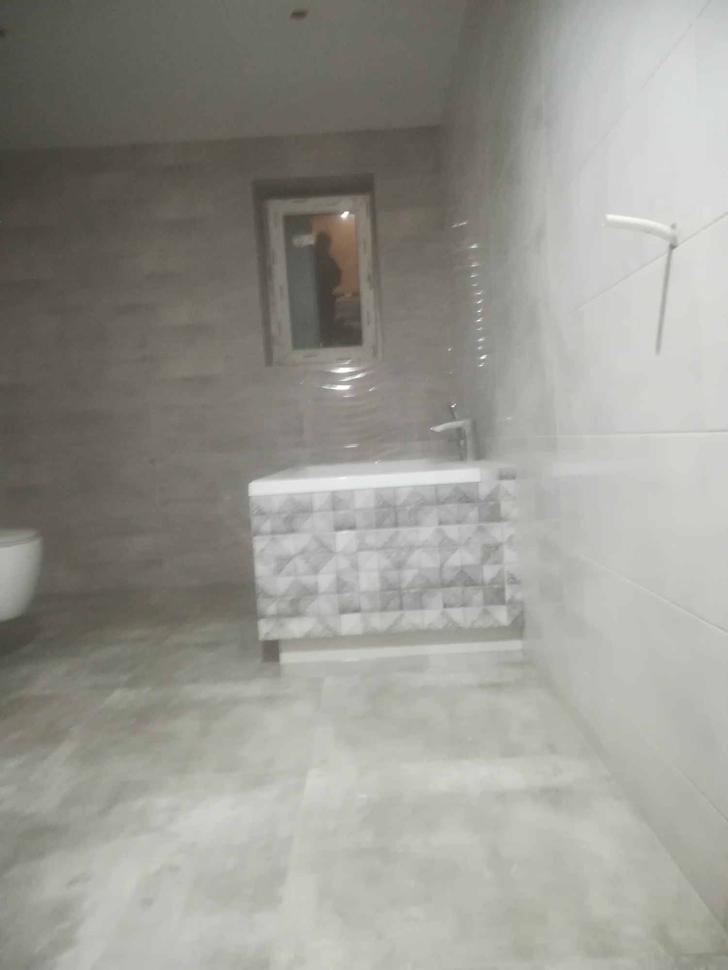 Wykończona łazienka z szarymi płytkami na podłodze i ścianach, umywalka z szafką w geometryczny wzór, widoczne okno z odbiciem sylwetki.