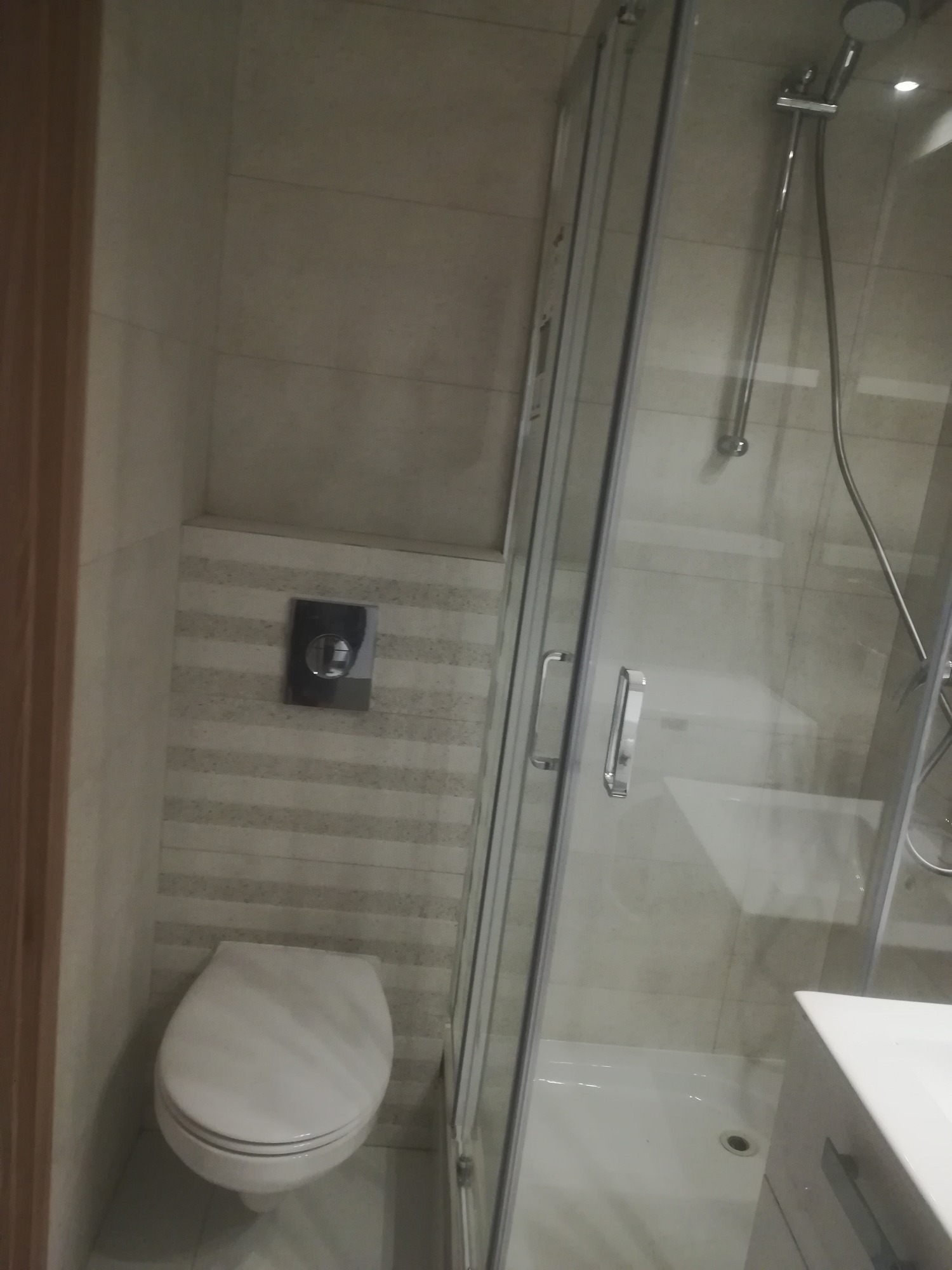 Wnętrze łazienki po remoncie: widok na toaletę podwieszaną z chromowanym przyciskiem spłukiwania, kabinę prysznicową z przesuwnymi drzwiami i fragment umywalki z szafką.