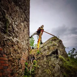 Pomiarowiec z niwelatorem na trójnogu, stojący na skalistej powierzchni obok starego, kamienno-ceglanego muru, pochmurne niebo w tle.