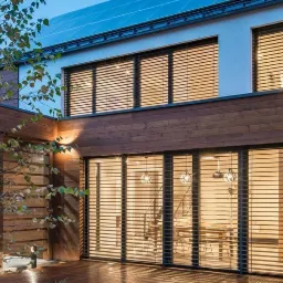 Nowoczesny dom z drewnianą elewacją i dużymi oknami z żaluzjami, widok z zewnątrz na oświetlony parter i piętro, pergola z samochodem w tle.