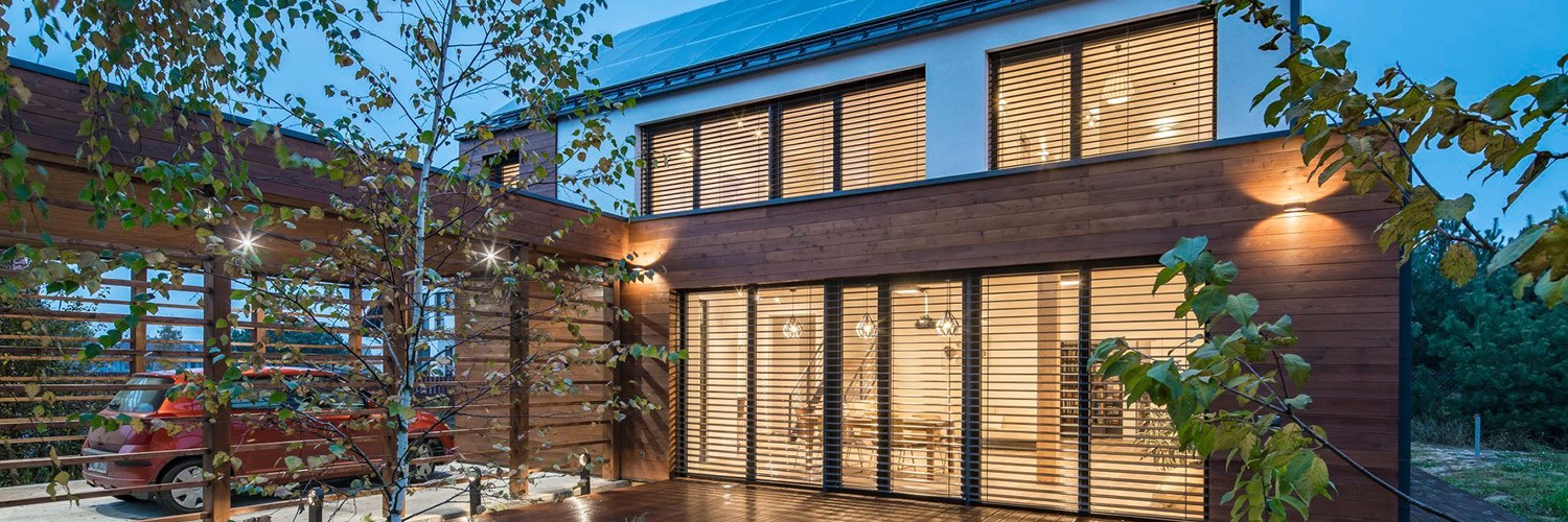 Nowoczesny dom z drewnianą elewacją i dużymi oknami z żaluzjami, widok z zewnątrz na oświetlony parter i piętro, pergola z samochodem w tle.