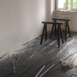 Przygotowane do malowania pomieszczenie z zabezpieczoną folią podłogą, dwoma kozłami malarskimi i wałkami do malowania w folii.