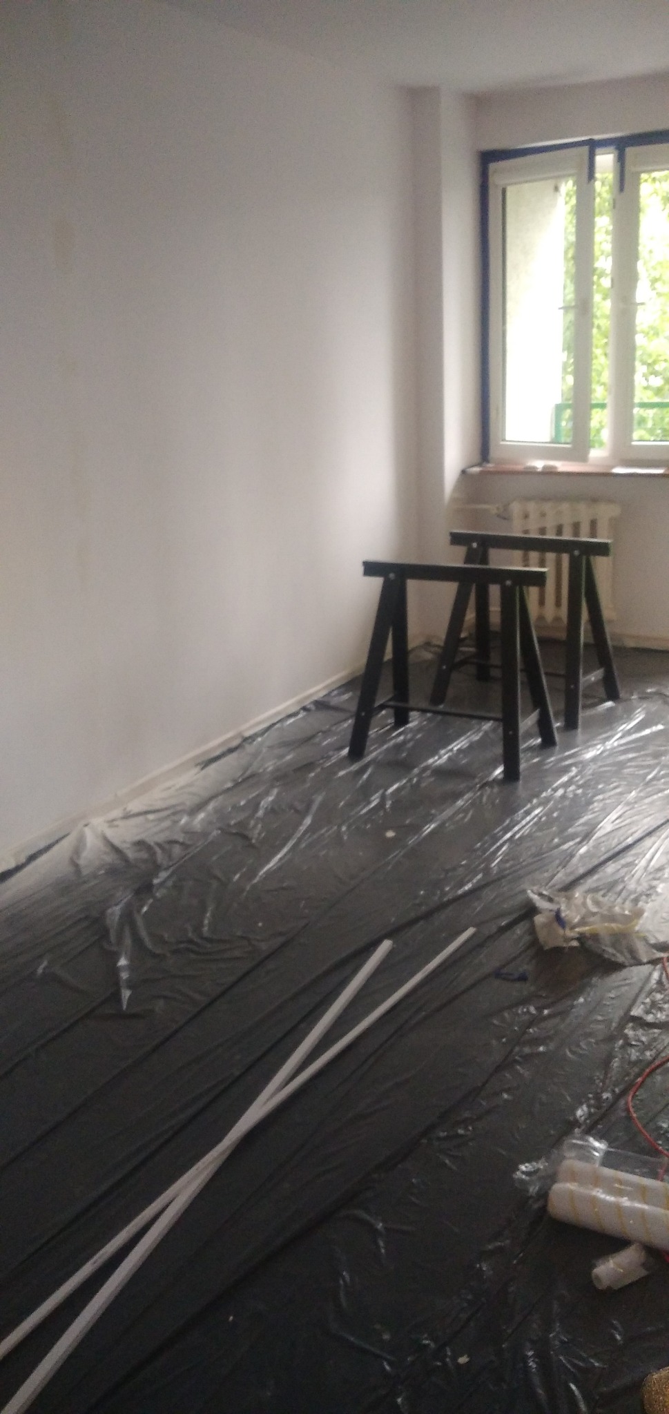 Przygotowane do malowania pomieszczenie z zabezpieczoną folią podłogą, dwoma kozłami malarskimi i wałkami do malowania w folii.