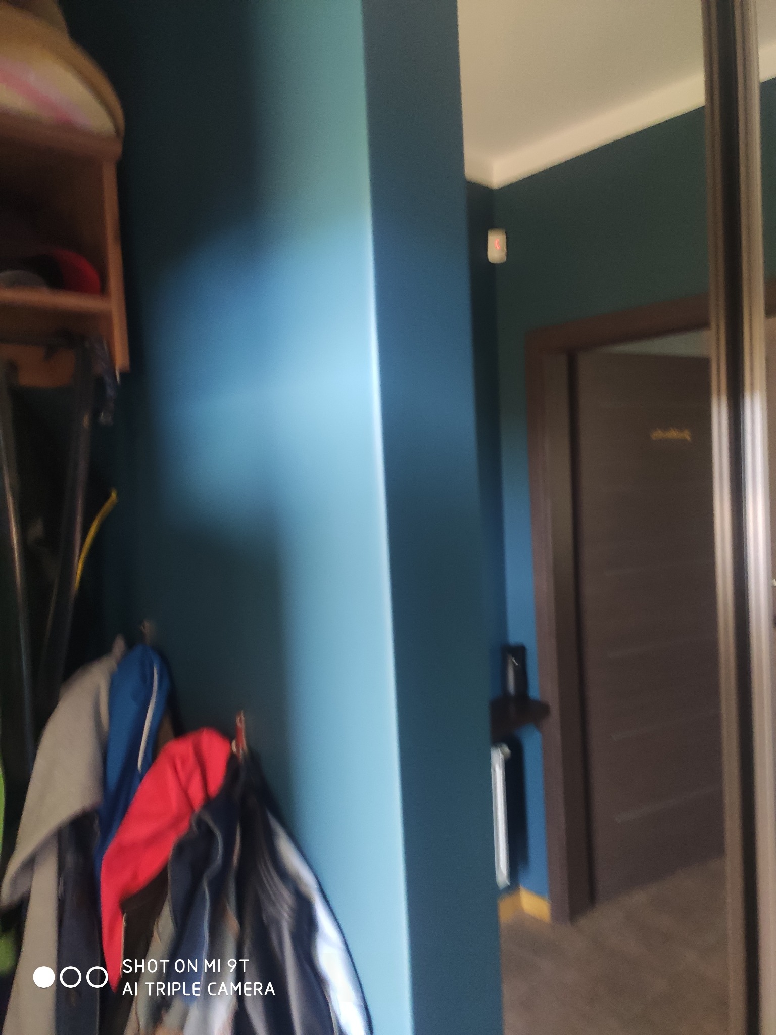 Fragment wnętrza mieszkania po remoncie, ściana pomalowana na niebiesko, widoczna część garderoby z ubraniami i fragment drzwi w kolorze ciemnego drewna.