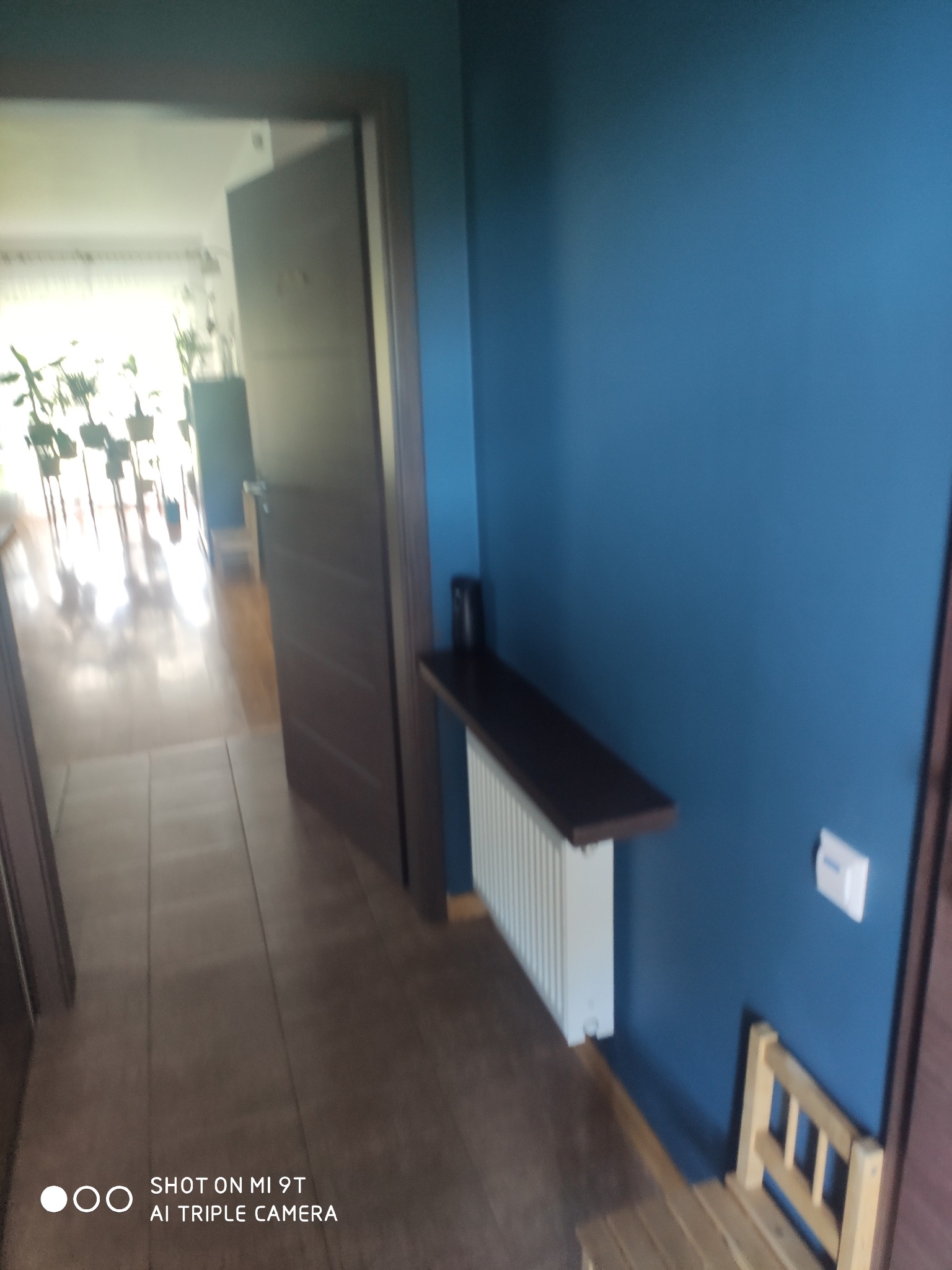 Niebieska ściana w korytarzu z ciemnymi drzwiami, grzejnikiem z półką i drewnianym krzesłem dla dziecka.