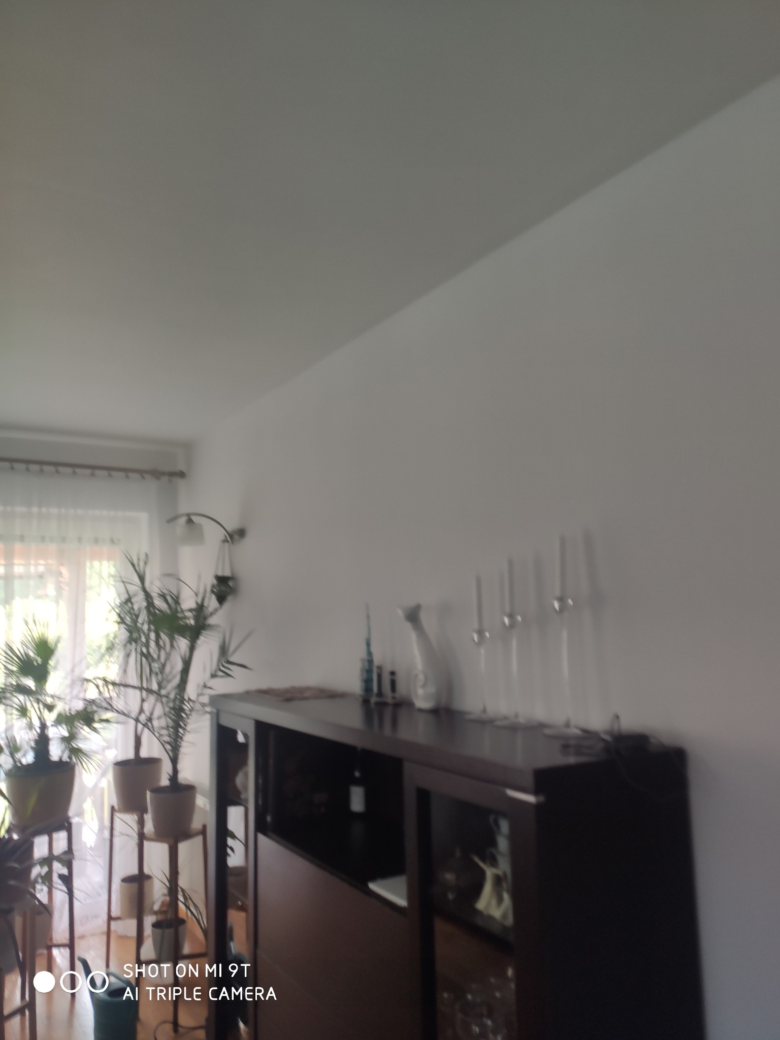 Jasne wnętrze pokoju z białymi ścianami, wyposażone w ciemnobrązową komodę z przeszklonymi drzwiczkami, na której stoją dekoracje i wysokie kieliszki. Po lewej stronie, przy oknie, widoczne rośliny...