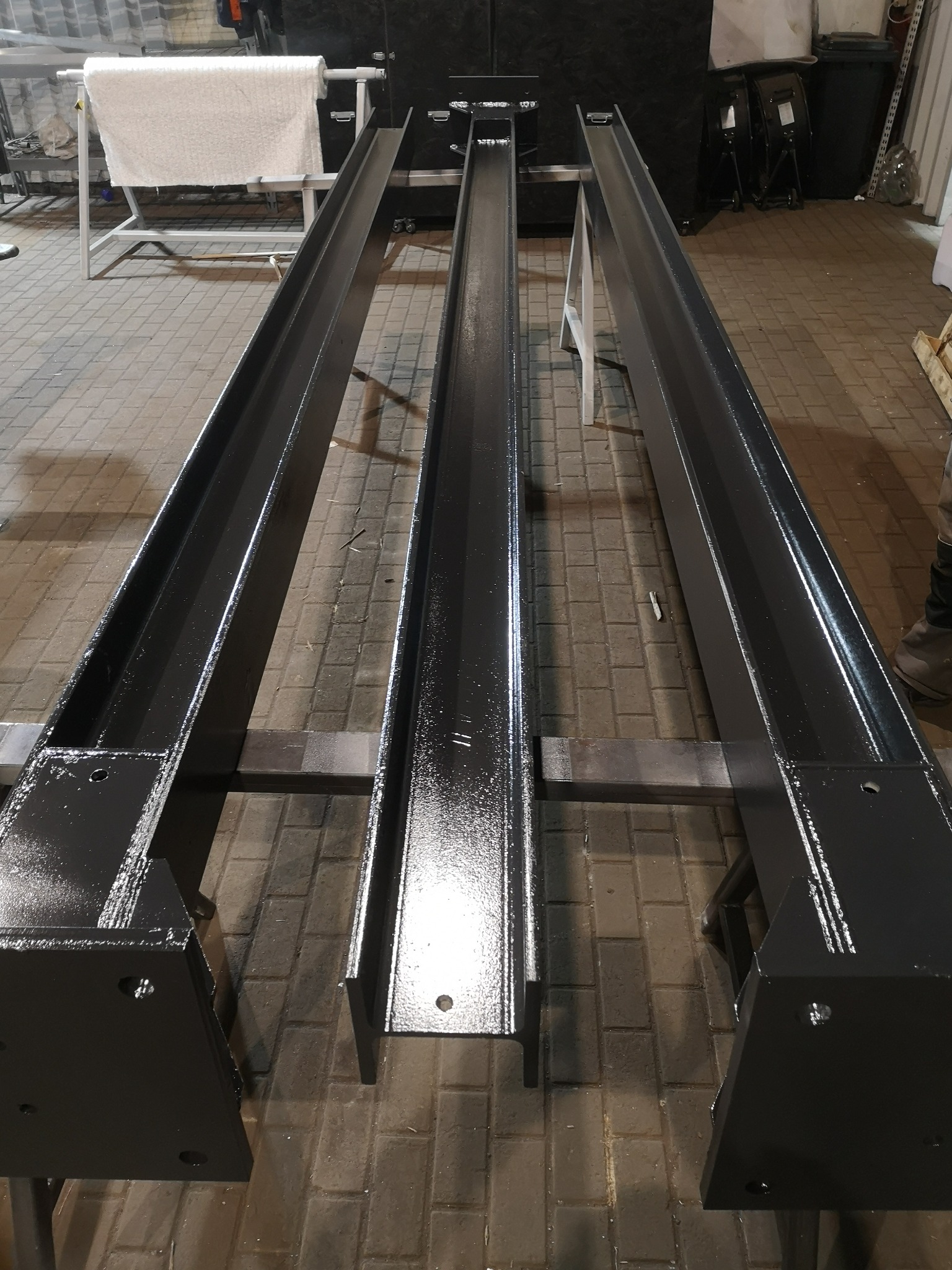 Trzy równoległe, pomalowane na czarno, stalowe belki H leżące na metalowych kozłach w warsztacie, widoczne ślady świeżego lakieru.